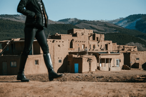 A person walks past traditional adobe buildings in a mountainous landscape.