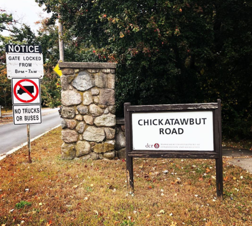 Sign for Chickatawbut Road with a notice about gate hours and restrictions on trucks and buses. Stone wall in background.