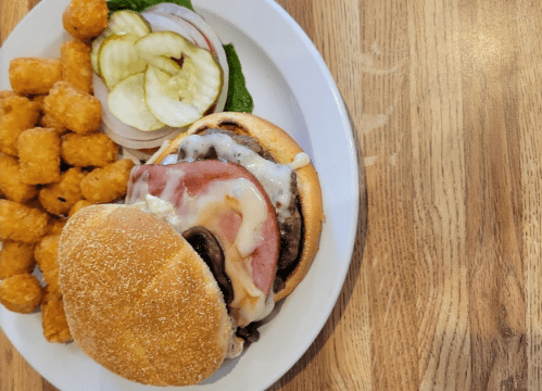 A plate with a cheeseburger topped with ham, pickles, and onions, alongside a serving of tater tots.