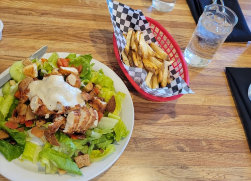 A plate of salad with grilled chicken, croutons, and dressing, alongside a basket of fries and a glass of water.