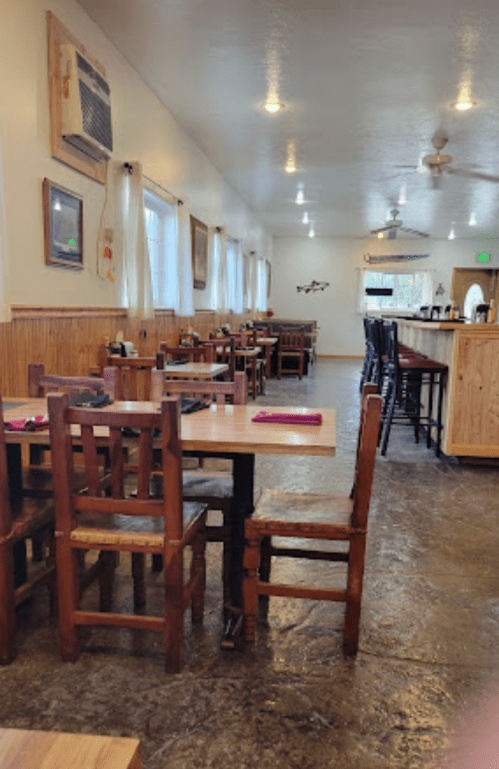 Interior of a restaurant with wooden tables and chairs, large windows, and a bar area in the background.