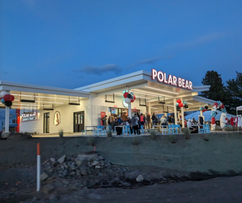 A modern ice cream shop named "Polar Bear" with outdoor seating and festive decorations, bustling with customers at dusk.