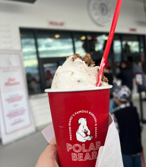 A hand holding a red cup of ice cream topped with cookie crumbles, in front of a burger and shake shop.