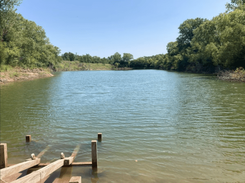 A calm lake surrounded by greenery, with a wooden dock extending into the water under a clear blue sky.