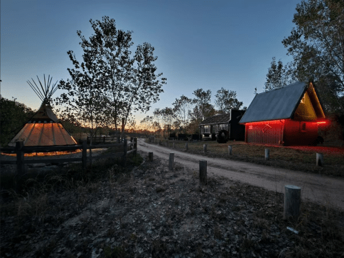A rustic scene at dusk featuring a teepee, a cabin with red lights, and a dirt path lined with wooden posts.