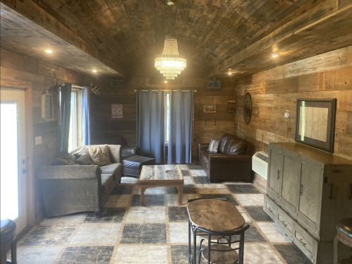Cozy living room with wooden walls, a chandelier, comfortable seating, and a checkered floor. Natural light from windows.