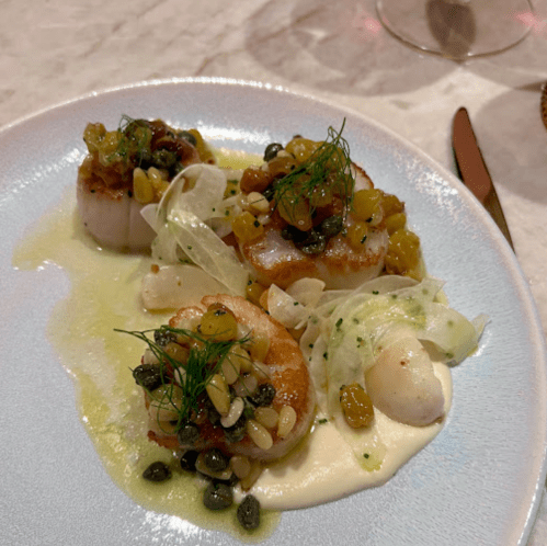 A plate of seared scallops topped with a mix of herbs, pine nuts, and raisins, served on a creamy sauce.