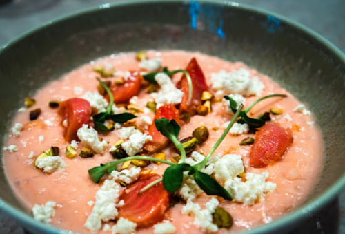A bowl of pink soup topped with cherry tomatoes, pistachios, and crumbled cheese, garnished with fresh herbs.
