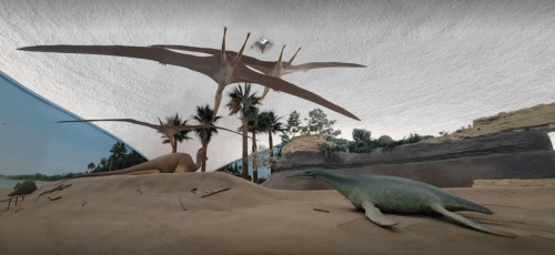 Dinosaur exhibit featuring pterosaurs flying above and a marine reptile on the ground in a sandy landscape.