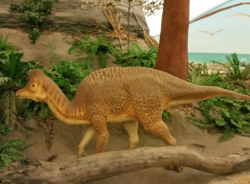 A dinosaur model walking through a prehistoric landscape with ferns and a beach in the background.