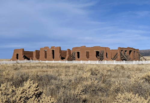 Historic adobe building with a white fence, set against a blue sky and dry grassland.