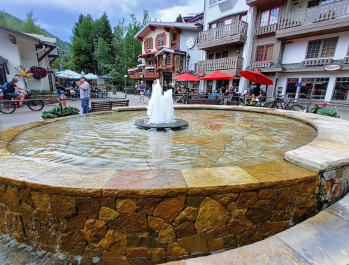 A stone fountain in a plaza surrounded by shops and outdoor seating, with people enjoying the area.