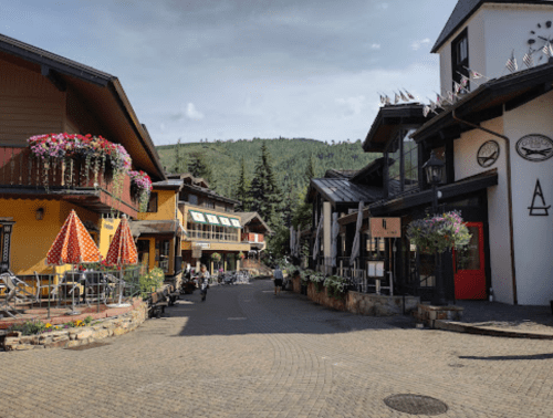 A charming street in a mountain town, featuring colorful buildings, outdoor seating, and lush greenery in the background.