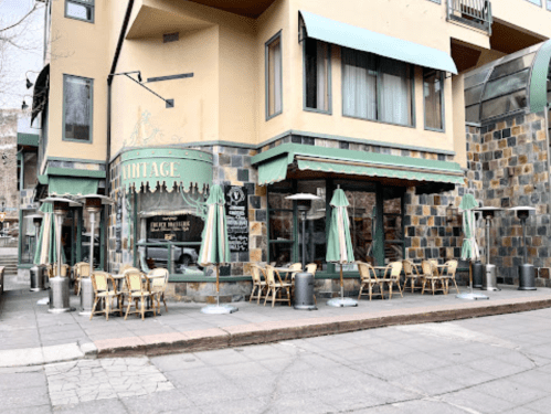 A quaint restaurant with outdoor seating, featuring green umbrellas and a stone facade, set in an urban environment.