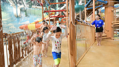 Children play and run through a water park, enjoying slides and splashes, with a staff member supervising nearby.
