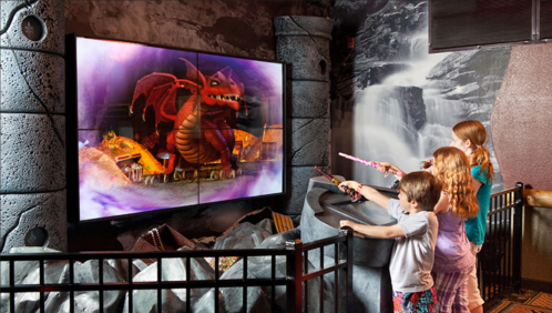 Children interact with a large screen displaying a dragon, using wands in a themed play area.