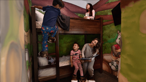 A cozy room with a bunk bed; a child climbs up while a mother reads to another child, with playful forest-themed decor.