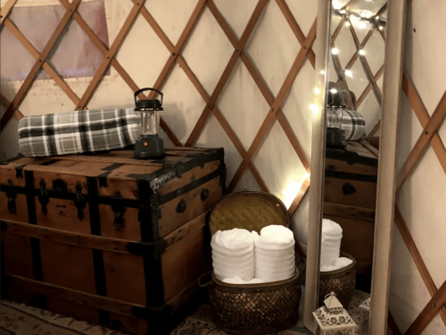 Cozy interior of a yurt featuring a wooden trunk, lantern, mirror, and neatly stacked towels in a basket.