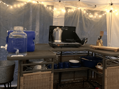A cozy outdoor kitchen setup with a camping stove, water jug, and various cooking utensils under string lights.