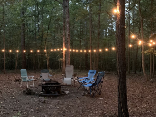 A cozy outdoor campsite with string lights, a fire pit, and several chairs surrounded by trees.