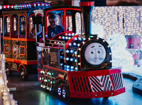 A colorful toy train with lights, featuring a smiling face, rides through a festive, illuminated setting.