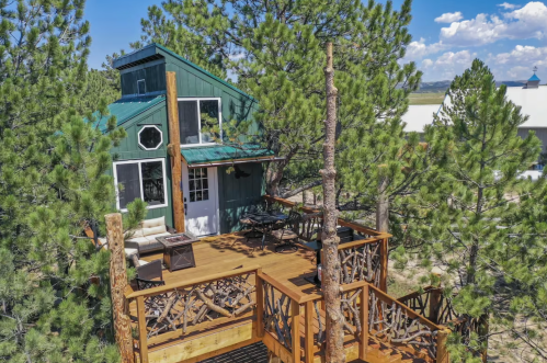 A cozy green cabin nestled among tall pine trees, featuring a wooden deck and outdoor seating.