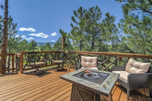 A wooden deck with seating and a fire pit, surrounded by tall trees and a scenic mountain view under a clear blue sky.