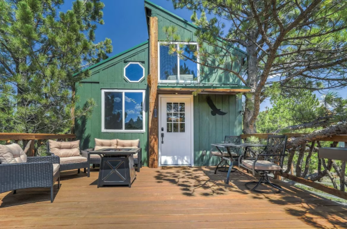 A green cabin surrounded by trees, featuring a deck with seating and a door leading inside.
