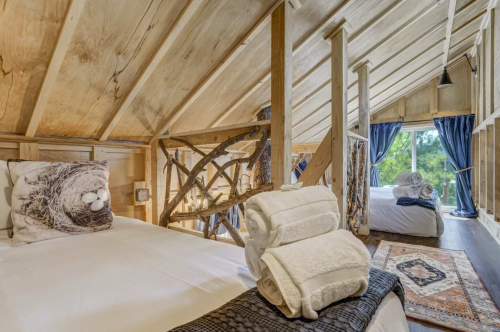 Cozy wooden cabin interior with two beds, decorative pillows, and neatly stacked towels, surrounded by nature.