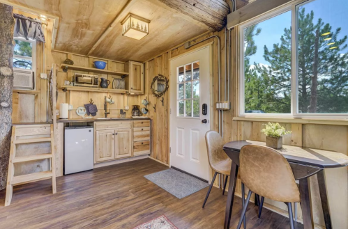 Cozy wooden interior of a small cabin with a kitchenette, dining table, and large windows showcasing nature outside.