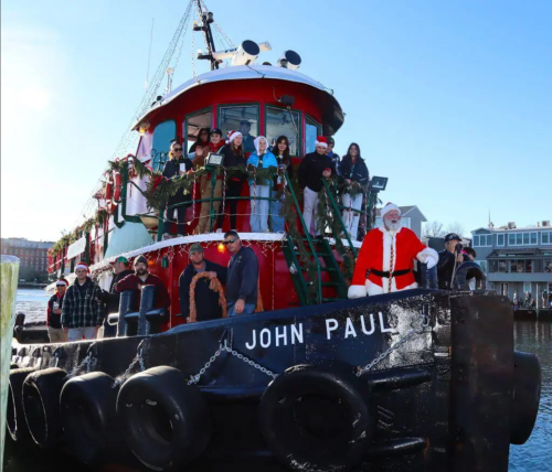 A festive group in Santa hats poses on a tugboat decorated for the holidays, with a Santa figure in front.