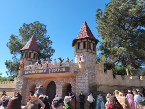 A stone castle with towers, featuring a "Renaissance Festival" sign, surrounded by a crowd in a festive atmosphere.