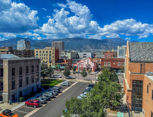 A cityscape view featuring buildings, parked cars, and mountains under a blue sky with fluffy clouds.