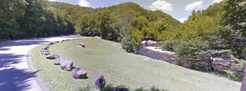 A scenic view of a grassy area by a river, surrounded by trees and mountains under a clear blue sky.