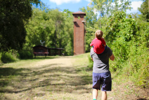 A person carries a child on their shoulders along a path in a wooded area, with a tall structure in the background.