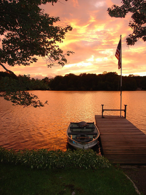 A serene lake at sunset, with vibrant orange skies, a small boat docked by a wooden pier, and an American flag.