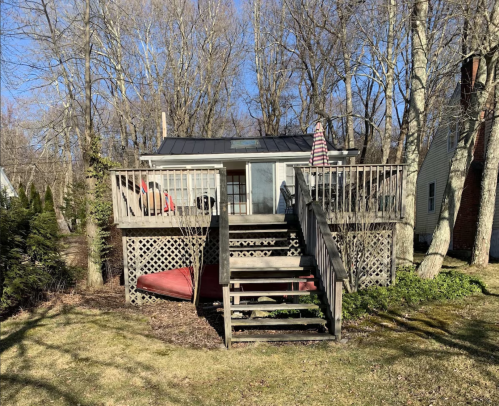 A small house with a deck and stairs, surrounded by trees and grass on a sunny day.