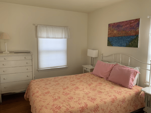 Cozy bedroom featuring a white bed with pink pillows, a dresser, and a colorful painting on the wall.
