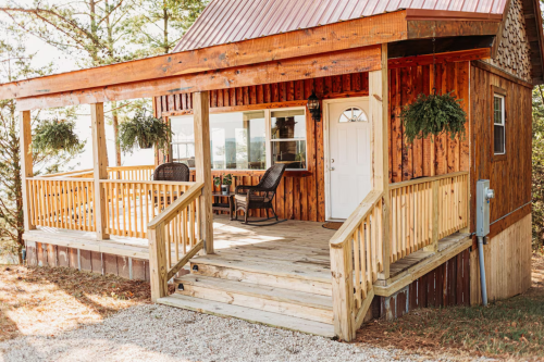 A cozy wooden cabin with a porch, featuring rocking chairs and greenery, set in a natural outdoor environment.