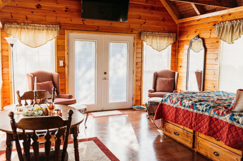 Cozy wooden cabin interior featuring a bed, two chairs, a dining table, and large windows with natural light.
