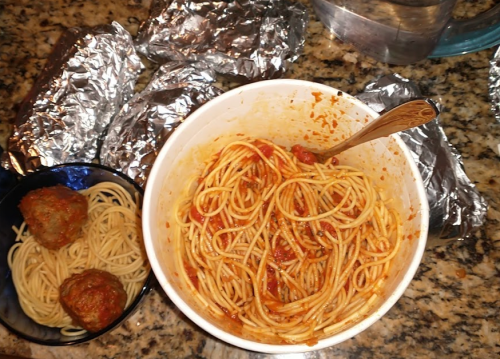 A bowl of spaghetti with meatballs, a side of meatballs, and several foil-wrapped items on a granite countertop.