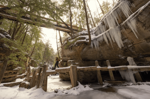 A snowy landscape with a wooden bridge, icy cliffs, and a person walking beneath a train trestle in a forested area.