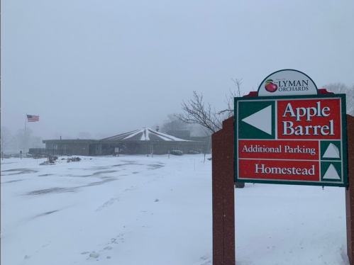 Snowy scene of Lyman Orchards' Apple Barrel sign, with a building in the background and an American flag waving.