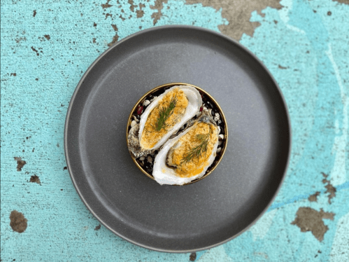 A gourmet dish featuring two oysters topped with a golden garnish, served on a dark plate against a textured turquoise background.