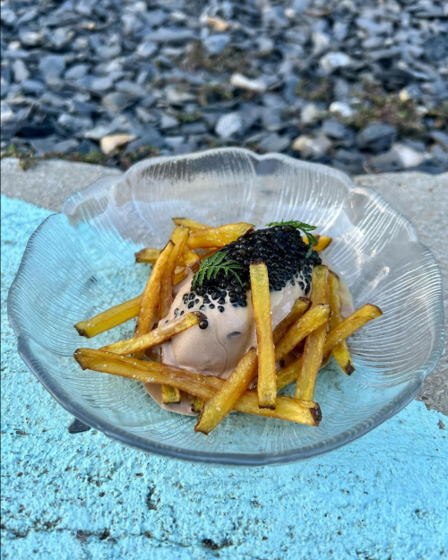A glass bowl filled with crispy fries topped with a scoop of creamy sauce and garnished with caviar and herbs.