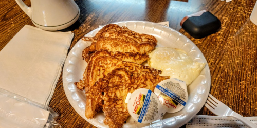 A plate of golden-brown French toast with a side of eggs and two containers of syrup on a wooden table.