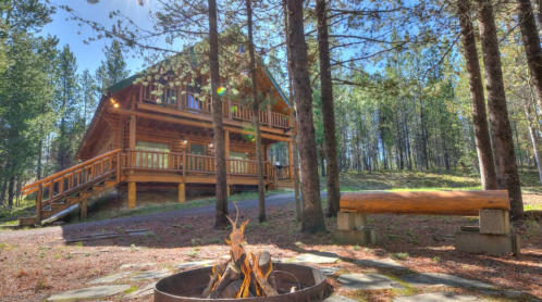 A cozy log cabin surrounded by trees, with a fire pit in the foreground and a bench nearby on a sunny day.
