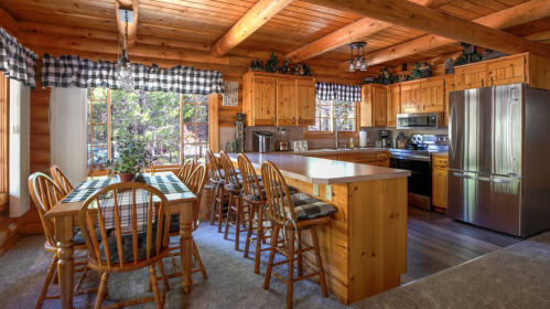 Cozy log cabin kitchen with wooden beams, a dining table, and modern appliances, surrounded by natural light.
