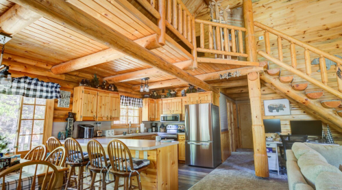 Cozy log cabin interior featuring a kitchen, dining area, and wooden staircase leading to a loft. Warm, rustic decor.