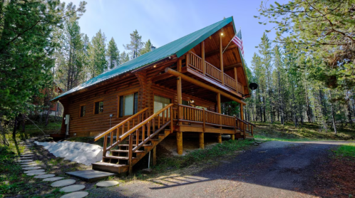 A wooden cabin with a green roof, surrounded by trees, featuring a porch and steps leading to the entrance.